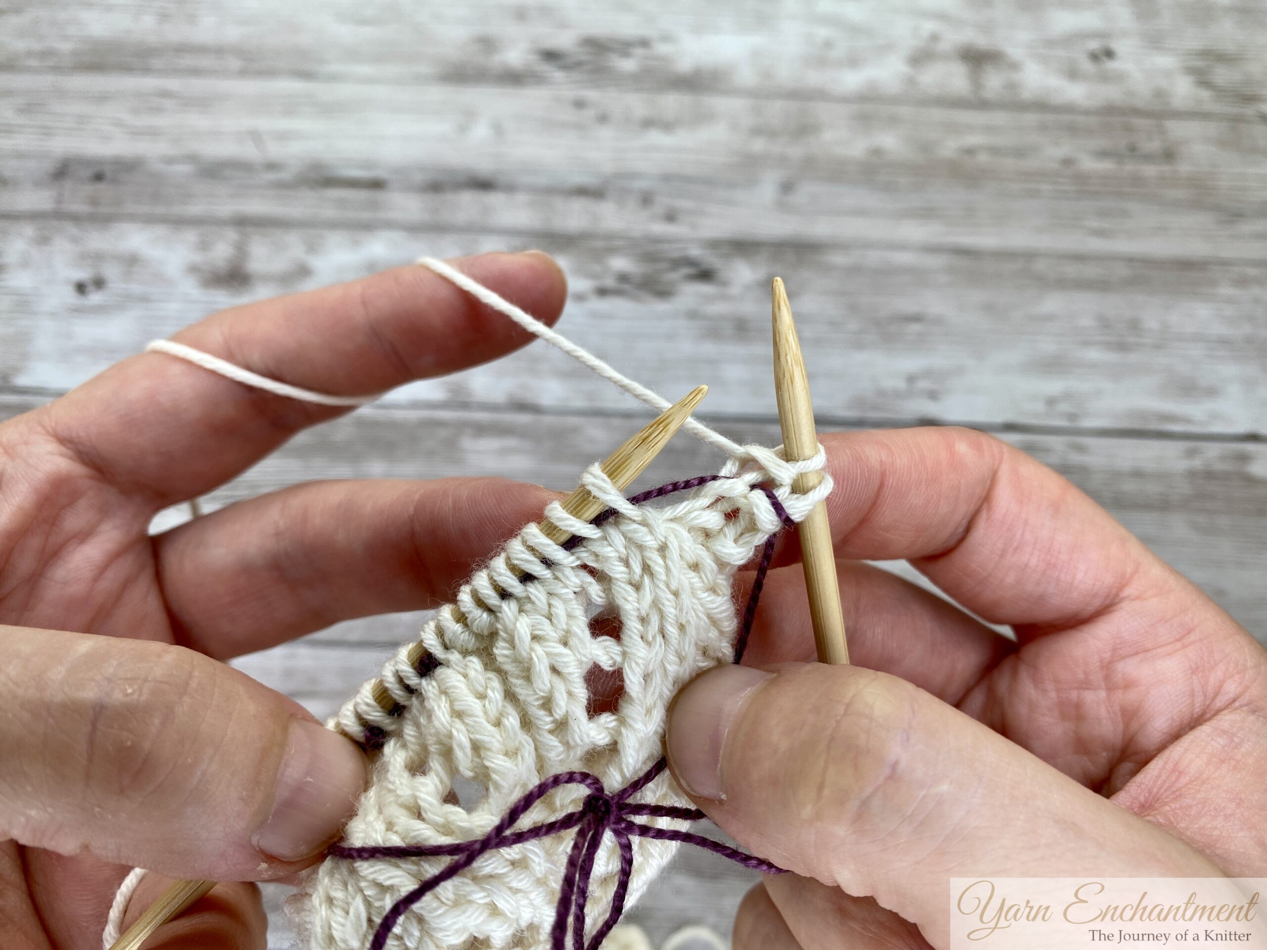 Hands knitting the next row with bamboo needles, purple lifeline sitting safely in the lace row below.