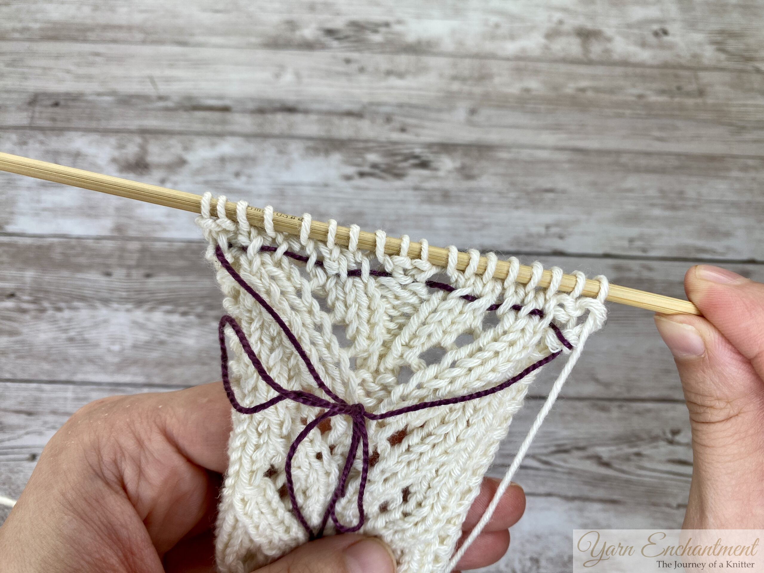 Finished purple lifeline running through a full row of cream lace knitting, live stitches still on the wooden needle.