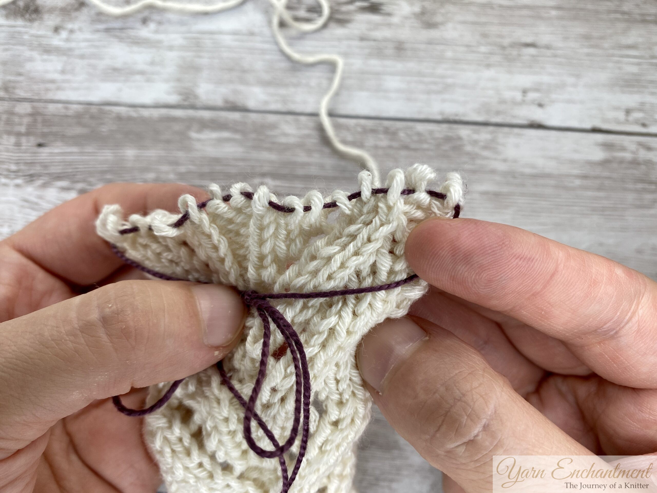 Hands holding cream lace knitting off the needles, purple lifeline threaded through the saved row of stitches.
