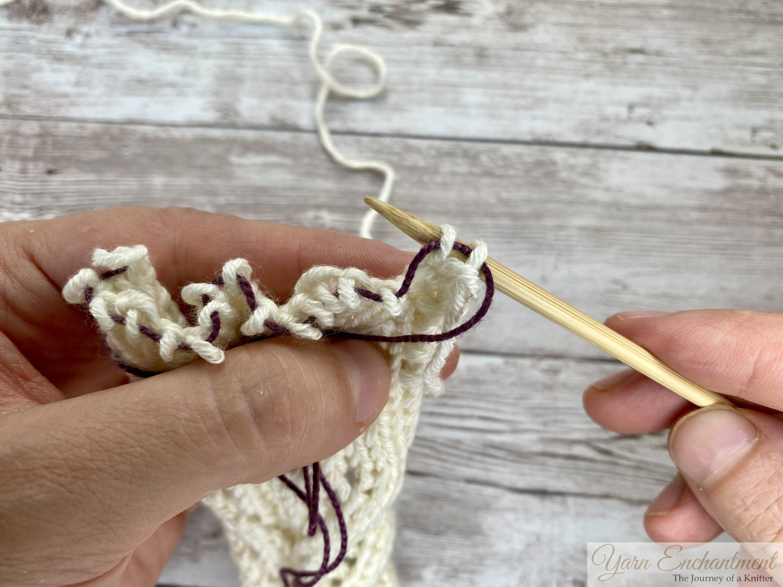 Cream lace knitting unraveling down to the purple lifeline, wooden needle ready to pick up the held stitches.