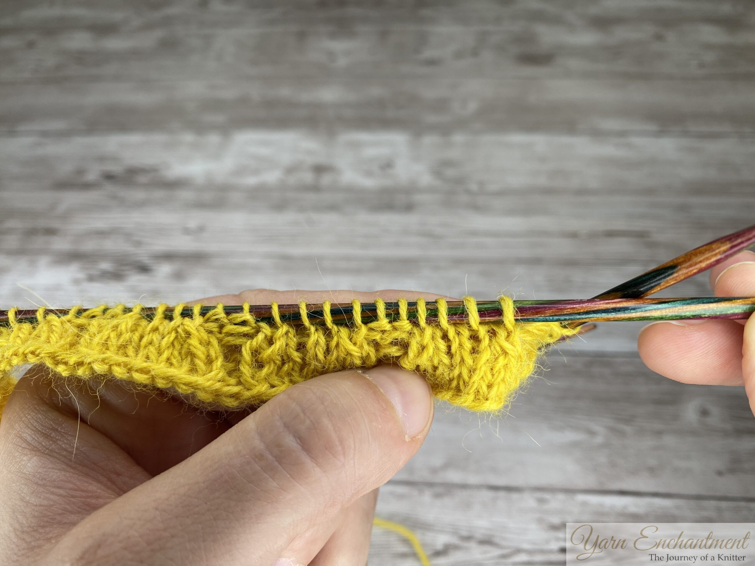 A close-up of two hands holding colorful metallic circular knitting needles, knitting with bright yellow yarn on a light grey wood-grain surface. The knitted fabric displays a knit 4, purl 2 ribbing pattern after a plain round. The right needle cable is printed with the text '3.50 mm'.