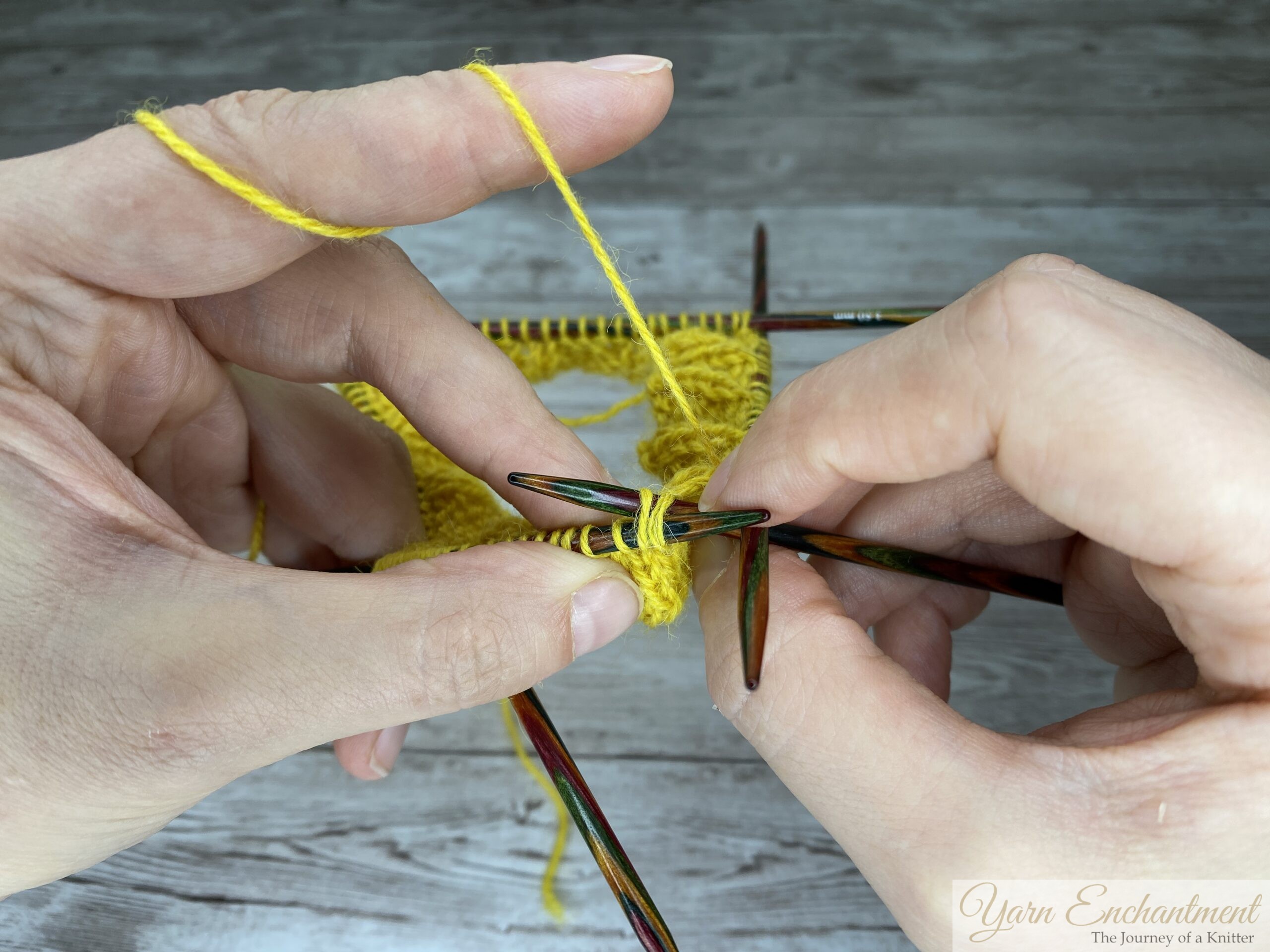 Hands holding colorful wooden circular knitting needles, working with bright yellow yarn. The right needle is inserted into the first two stitches on the left needle to begin the decrease round (SSK). The background is a light grey wood-grain surface.