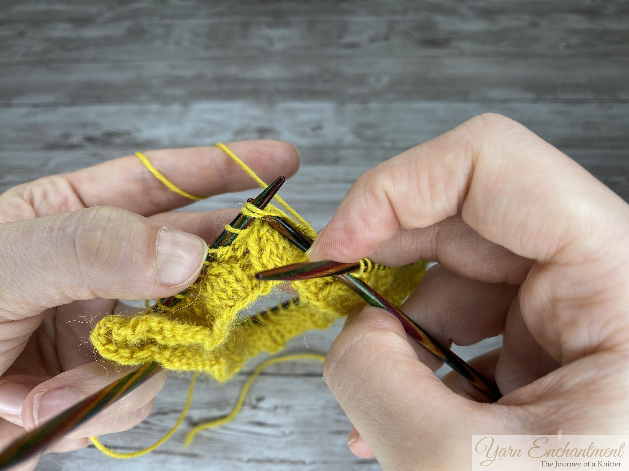 A close-up of the knitting process showing the right needle positioned through two stitches on the left needle to execute the decrease. The left hand holds the yellow working yarn taut, and the knitted fabric shows a K4, P2 ribbing pattern.