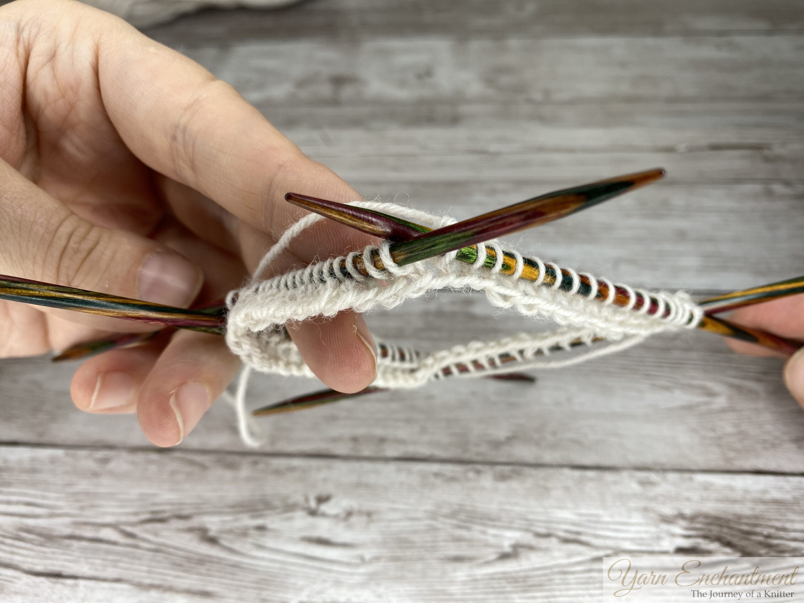 A knitter's hands holding colorful wooden circular needles with white yarn, resting against a light grey wood-grain background.