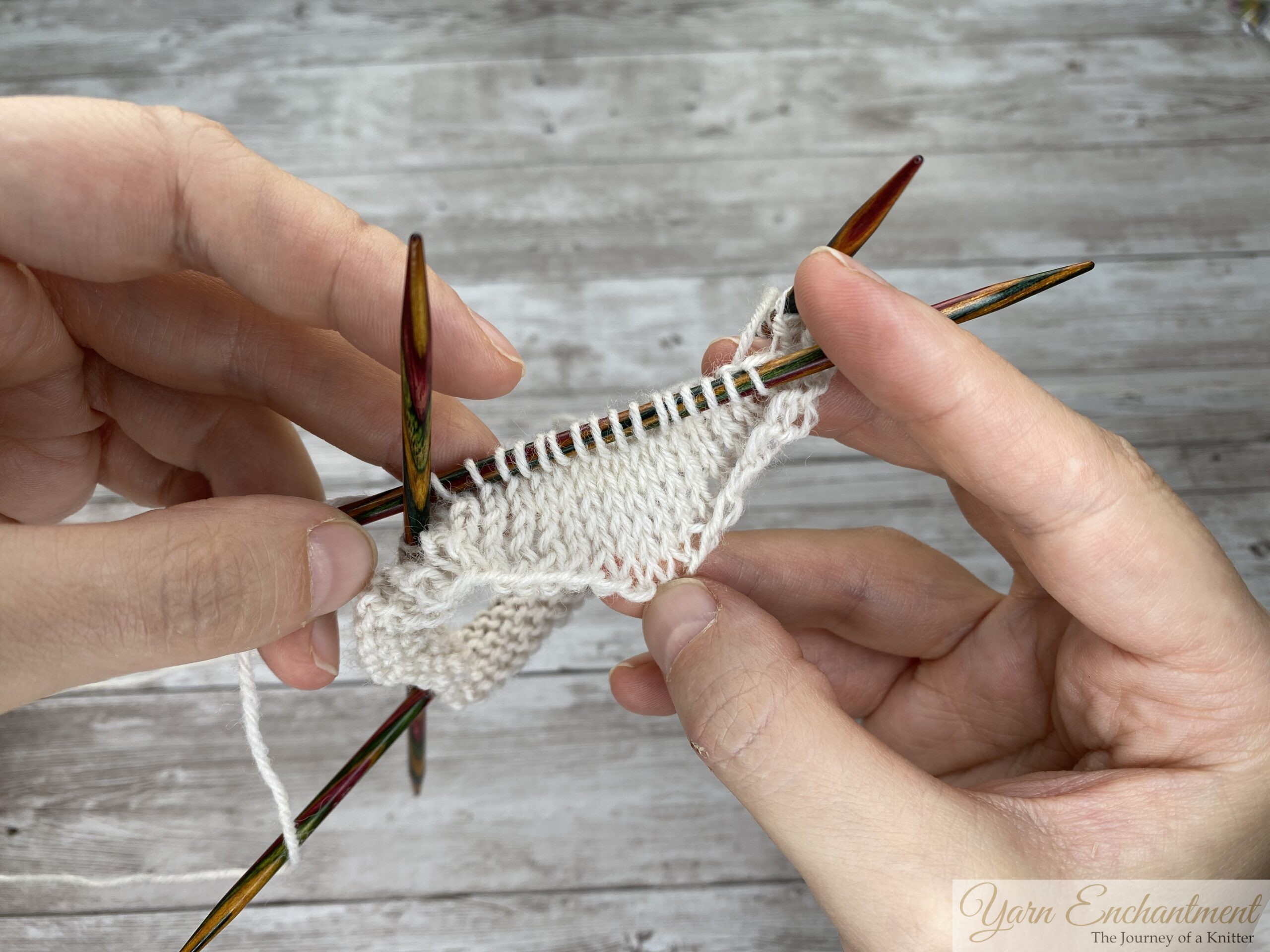 Close-up of white yarn stitches on colorful circular needles being held by hands over a grey wood surface.