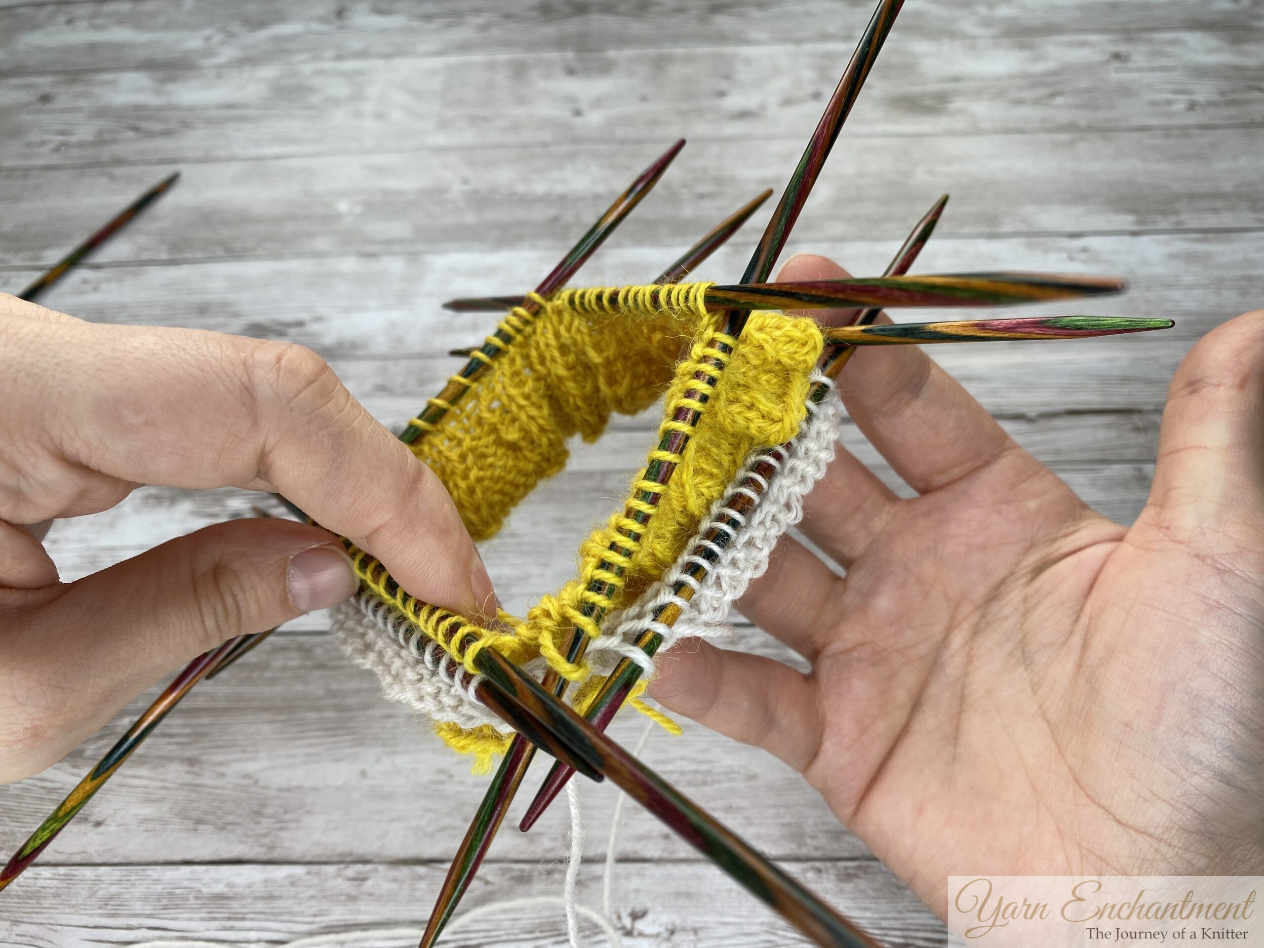 A close-up of a knitting project on colorful wooden double-pointed needles. The knitter's hands are adjusting the needles, showing the bright yellow ruffle alongside the newly cast-on white sock yarn against a grey wood-grain surface.