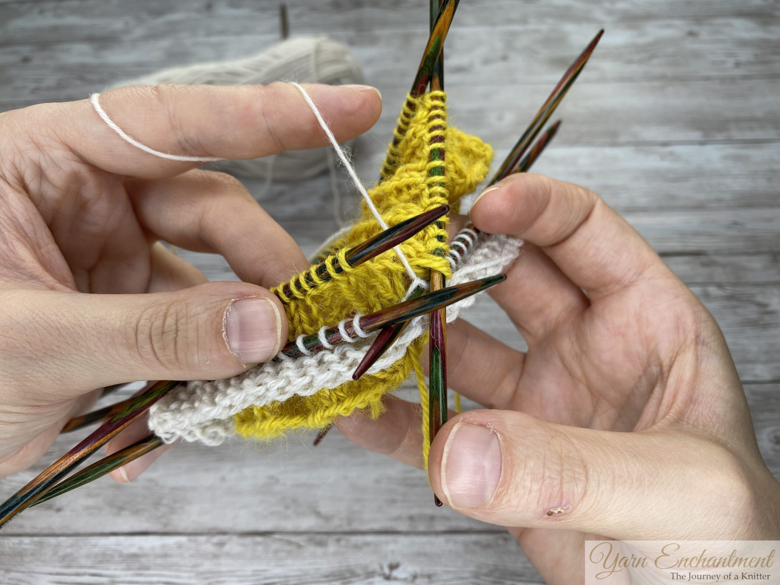 A close-up of a knitter's hands holding colorful wooden double-pointed needles. White yarn is being actively worked into the edge of the textured yellow ruffle, with the ball of white yarn resting in the background against a light grey wood-grain surface.