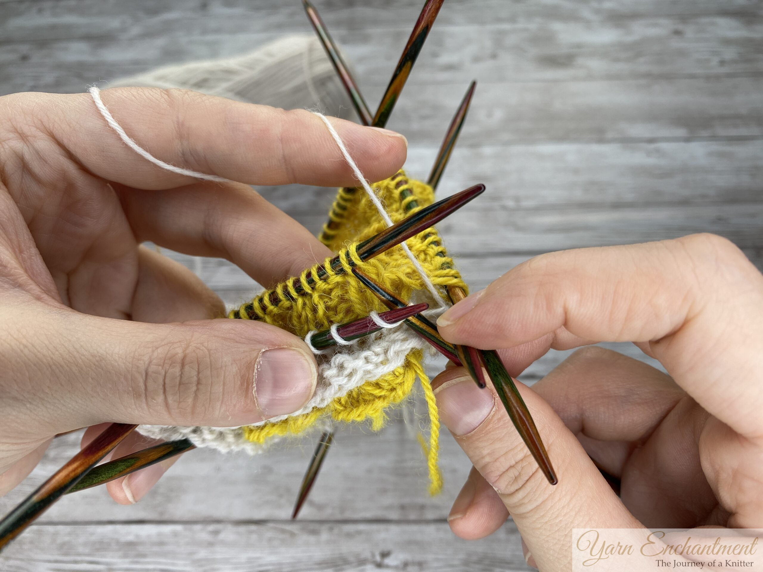 A detailed view of a knitting project on colorful wooden double-pointed needles. The knitter's hands are manipulating the needles to knit white yarn into the textured yellow ruffle section, set against a grey wood-grain background.