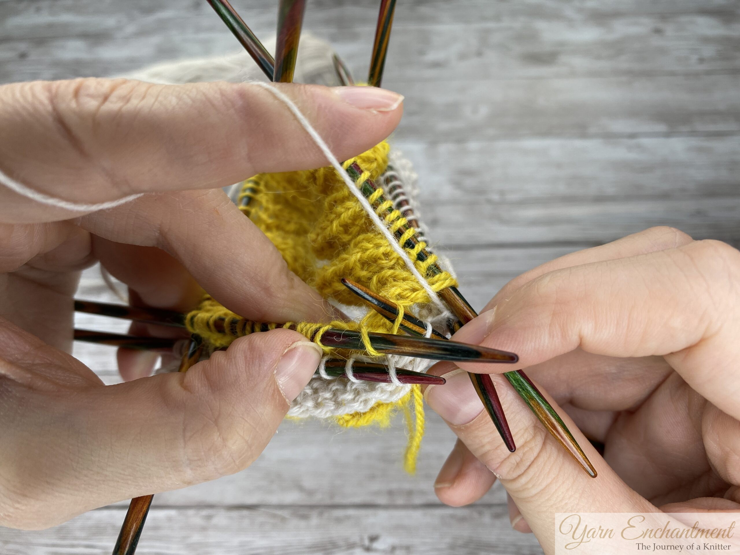 A tight close-up of a knitting project in progress. Colorful wooden double-pointed needles hold both the yellow ruffle and the white sock yarn. The right needle is wrapped with white yarn, preparing to pull through a stitch, over a grey wood-grain surface.
