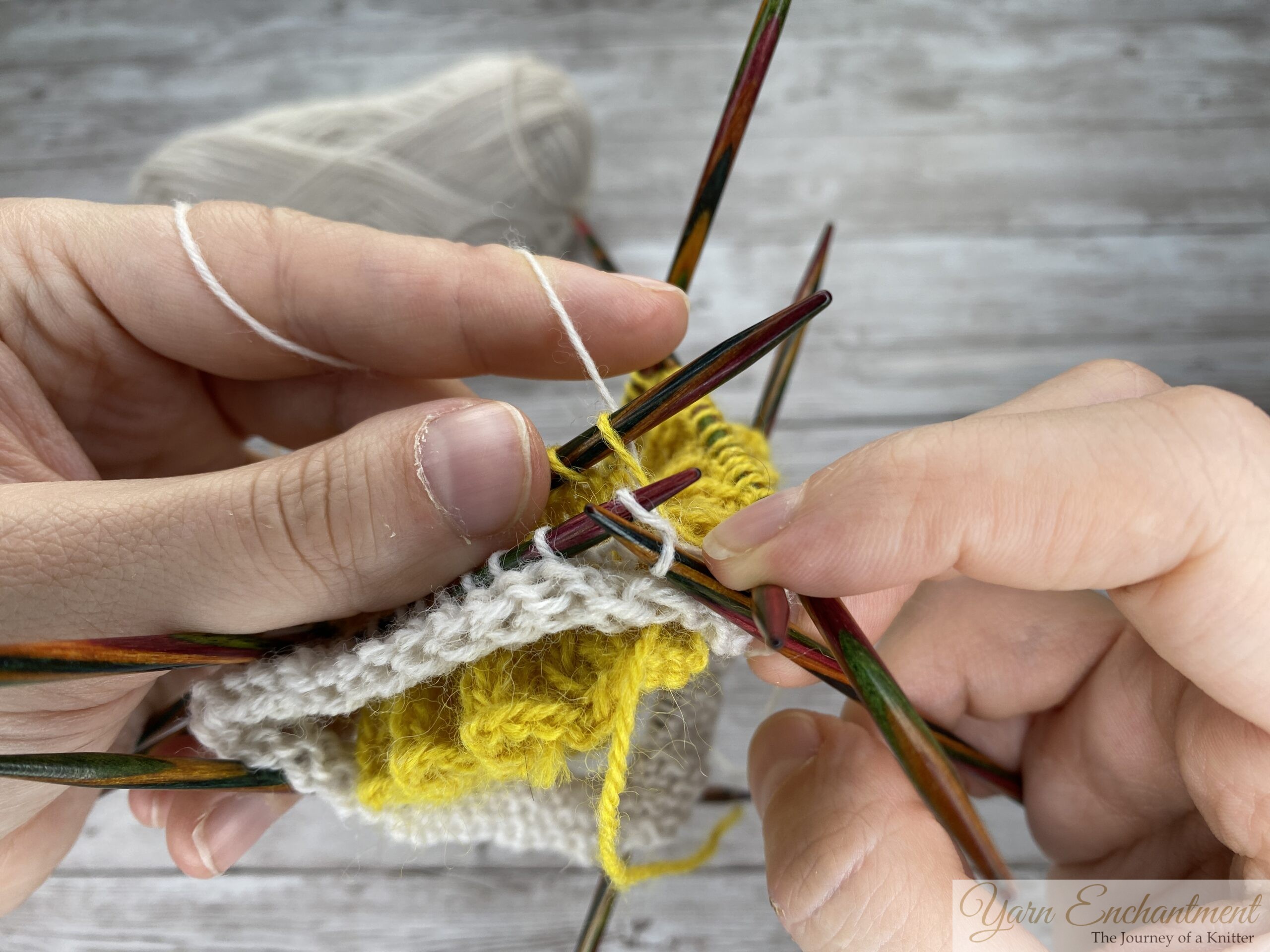 A detailed view of a knitting project on several colorful wooden double-pointed needles. The knitter's hands are actively wrapping and pulling the white working yarn through stitches to join the white sock section to the yellow ruffle, set against a grey wood-grain background.
