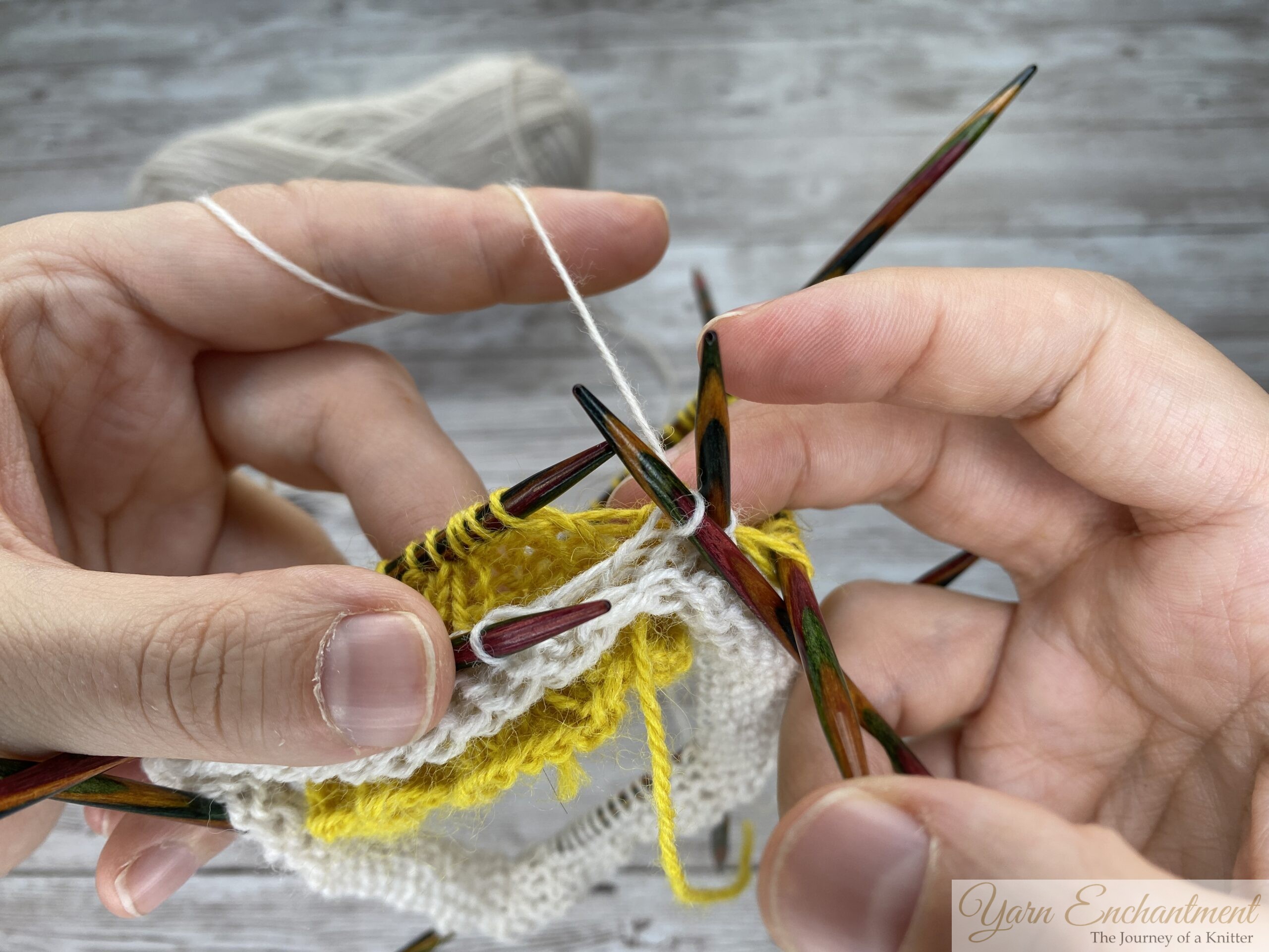 A close-up of a knitter's hands holding multiple colorful wooden double-pointed needles. White working yarn is tensioned over the left index finger and is being actively knit into the edge of the textured yellow ruffle. A ball of white yarn rests in the blurry background against a light grey wood-grain surface.
