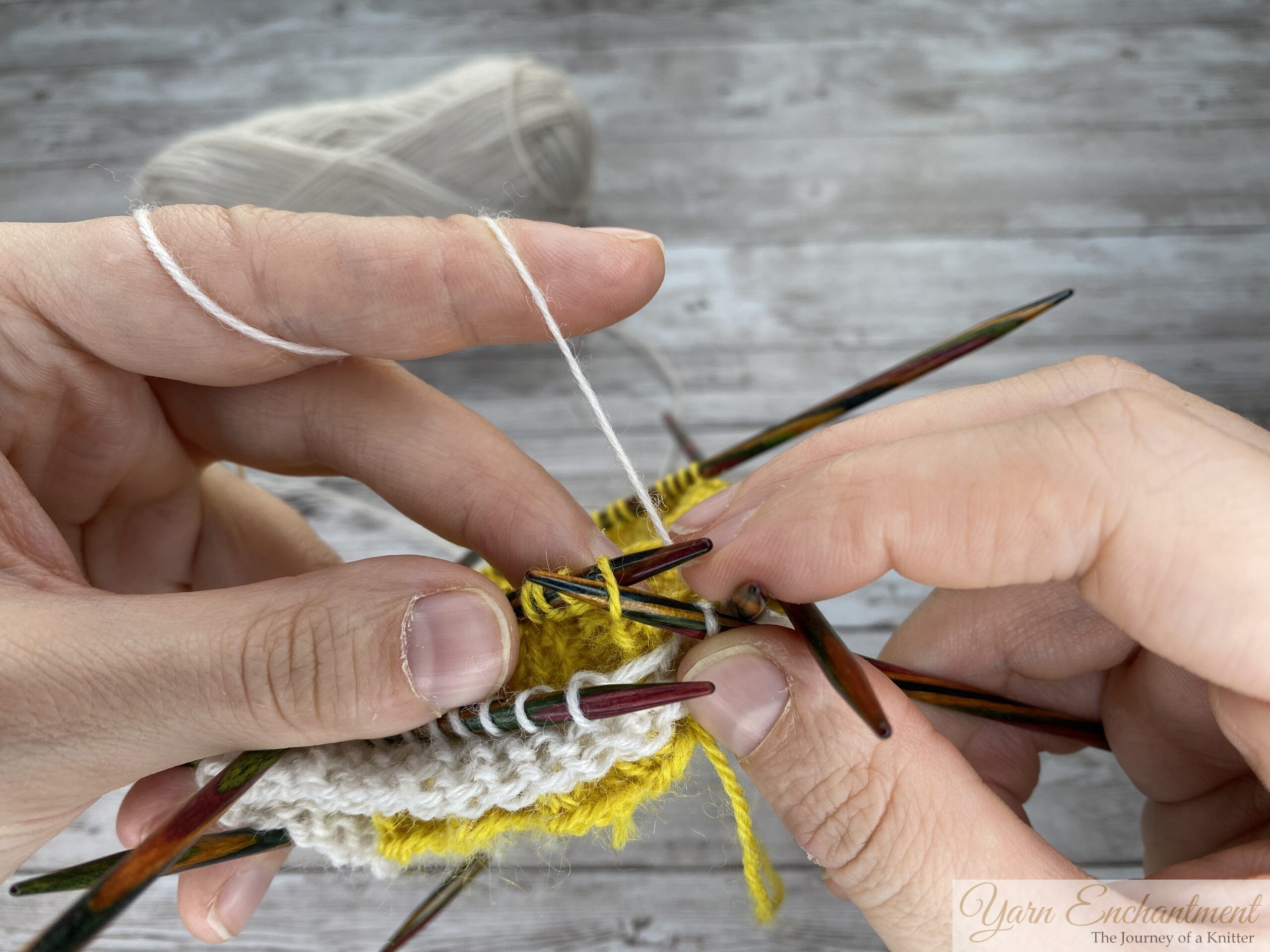 A close-up of a knitter's hands holding multiple colorful wooden double-pointed needles. White working yarn is tensioned over the left index finger and is being actively knit into the edge of the textured yellow ruffle. A ball of white yarn rests in the blurry background against a light grey wood-grain surface.