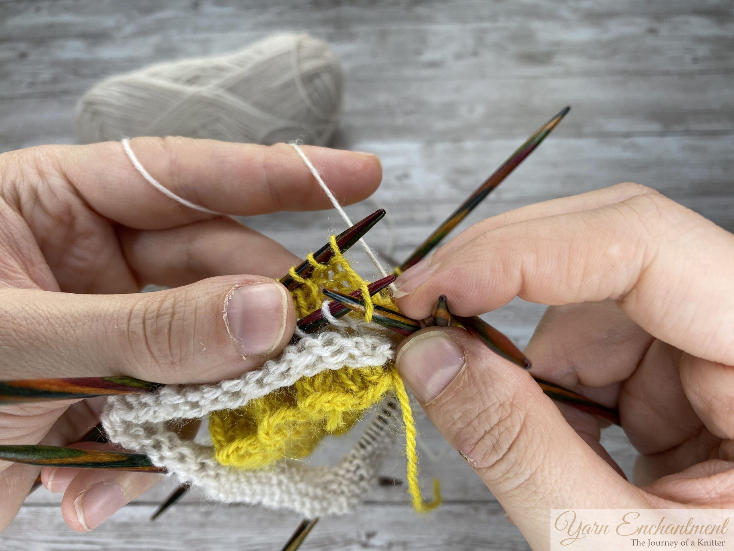 A detailed view of a knitting project on several colorful wooden double-pointed needles. The knitter's hands are actively wrapping and pulling the white working yarn through stitches to join the white sock section to the yellow ruffle, set against a grey wood-grain background.