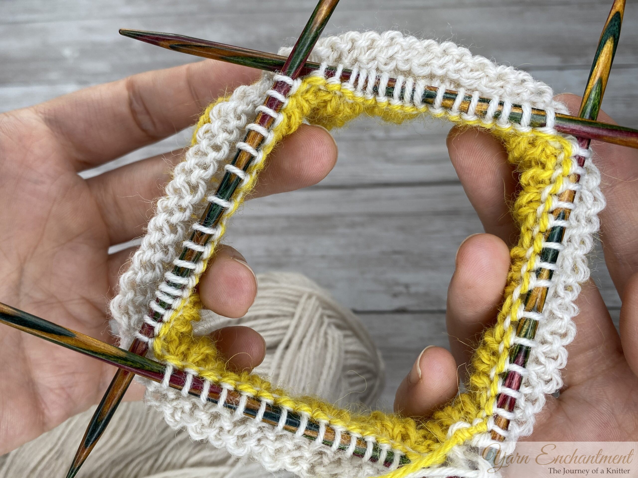 A top-down view of a circular knitting project on colorful wooden double-pointed needles. The work shows the yellow ruffle neatly joined to a round of white yarn, with another yellow section forming the base of the sock. A ball of white yarn rests in the background against a light grey wood-grain surface.