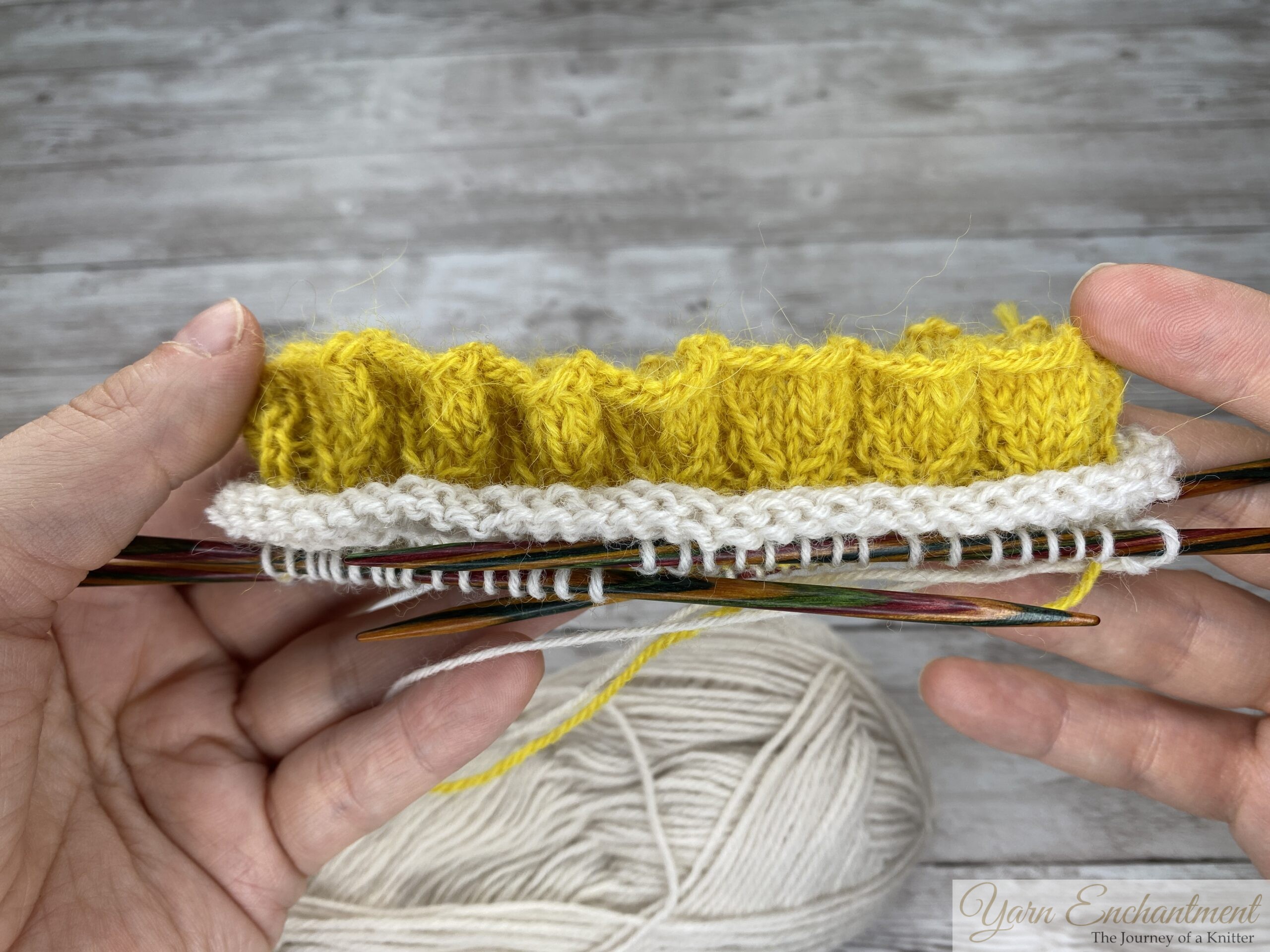 A close-up of a knitting project held in two hands. The project shows the textured yellow ruffle neatly attached to a smooth white knitted band on colorful wooden double-pointed needles. A large ball of white yarn sits in the foreground against a light grey wood-grain surface.