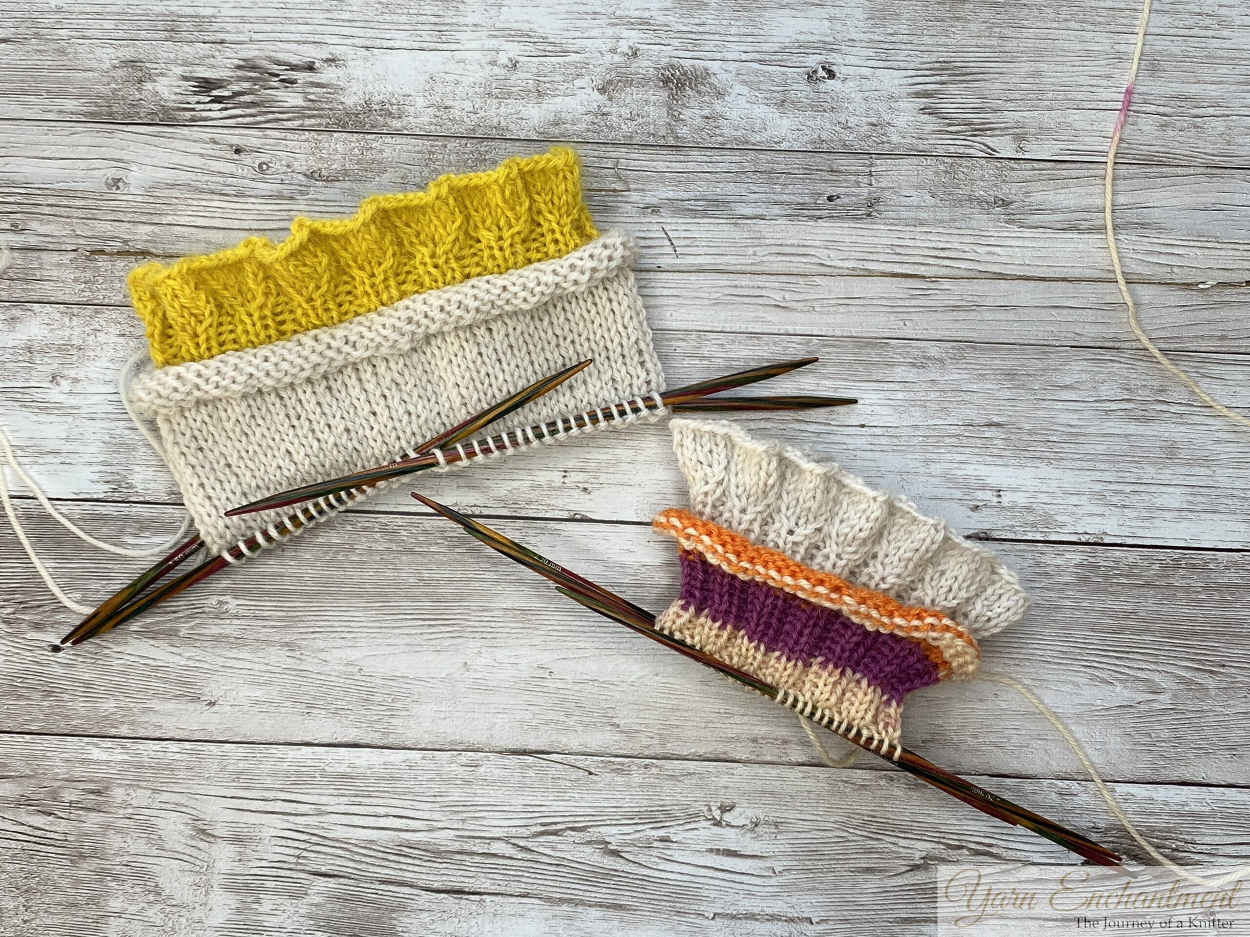 A flat lay of two knitting projects in progress, resting on a white-washed wood-grain table. On the top left, a project features a bright yellow ruffled edge joined to a white stockinette sock section on colorful wooden double-pointed needles. On the bottom right, another project features a white ruffle, a thin orange stripe, a wide purple stripe, and a tan section, also on colorful wooden double-pointed needles.
